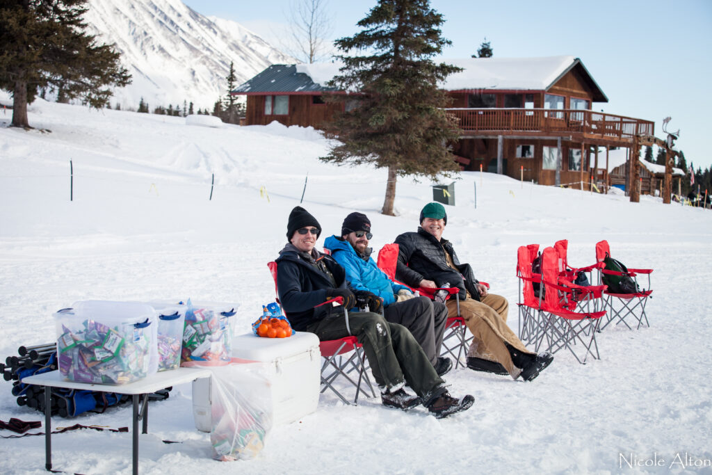 A group of male pilots sit on the sidelines enjoying the events during Iditarod at Rainy Pass checkpoint