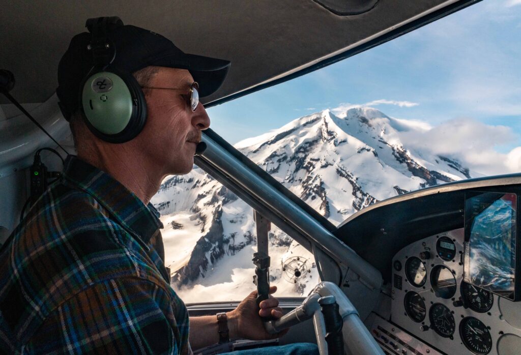 Male pilot navigates rugged terrain with volcanic Mt Redoubt seen from the window